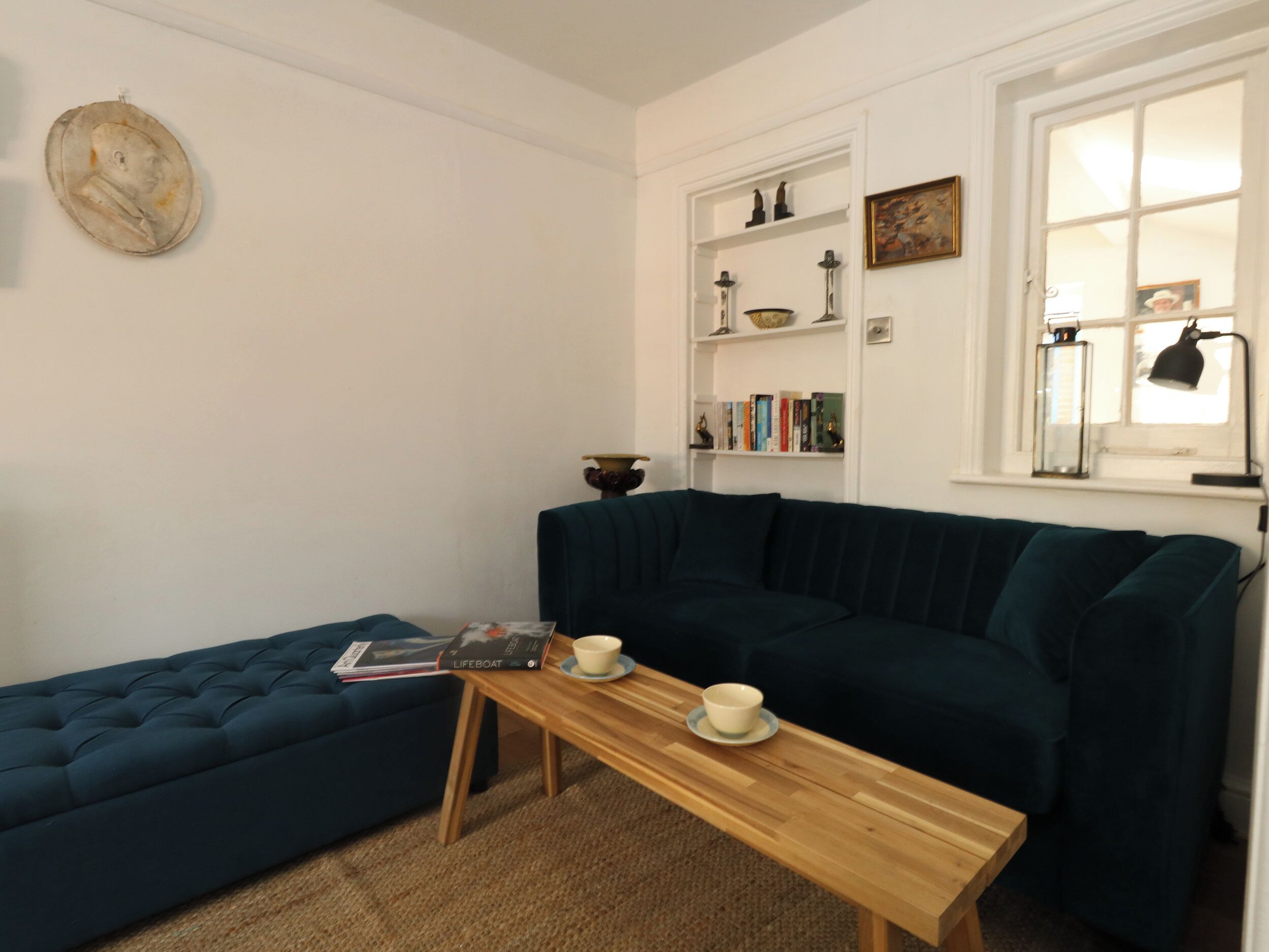 Una sala de estar acogedora con un sofá azul oscuro, una mesa de centro de madera con una revista y una taza, y una estantería con diversos objetos.