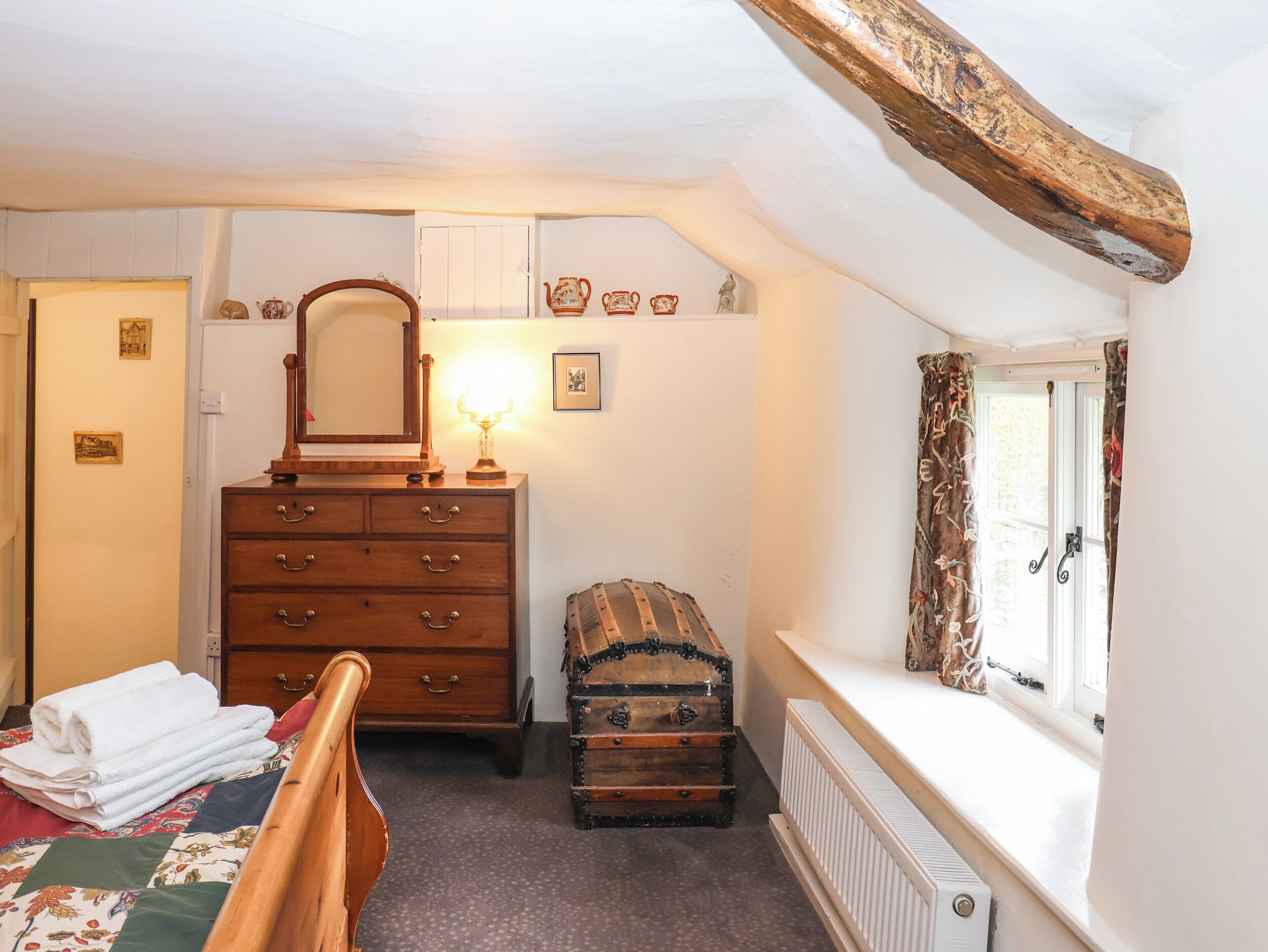 Ein Schlafzimmer mit einem Holzkoffer, einem Spiegel, einer Lampe, einem Truhenkasten und einem Fenster mit Vorhängen.