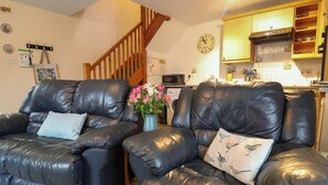 A living room with two black leather sofas, a wooden staircase, a kitchen with wooden cabinets, and a wall clock. - Penny Corner (Bridport)