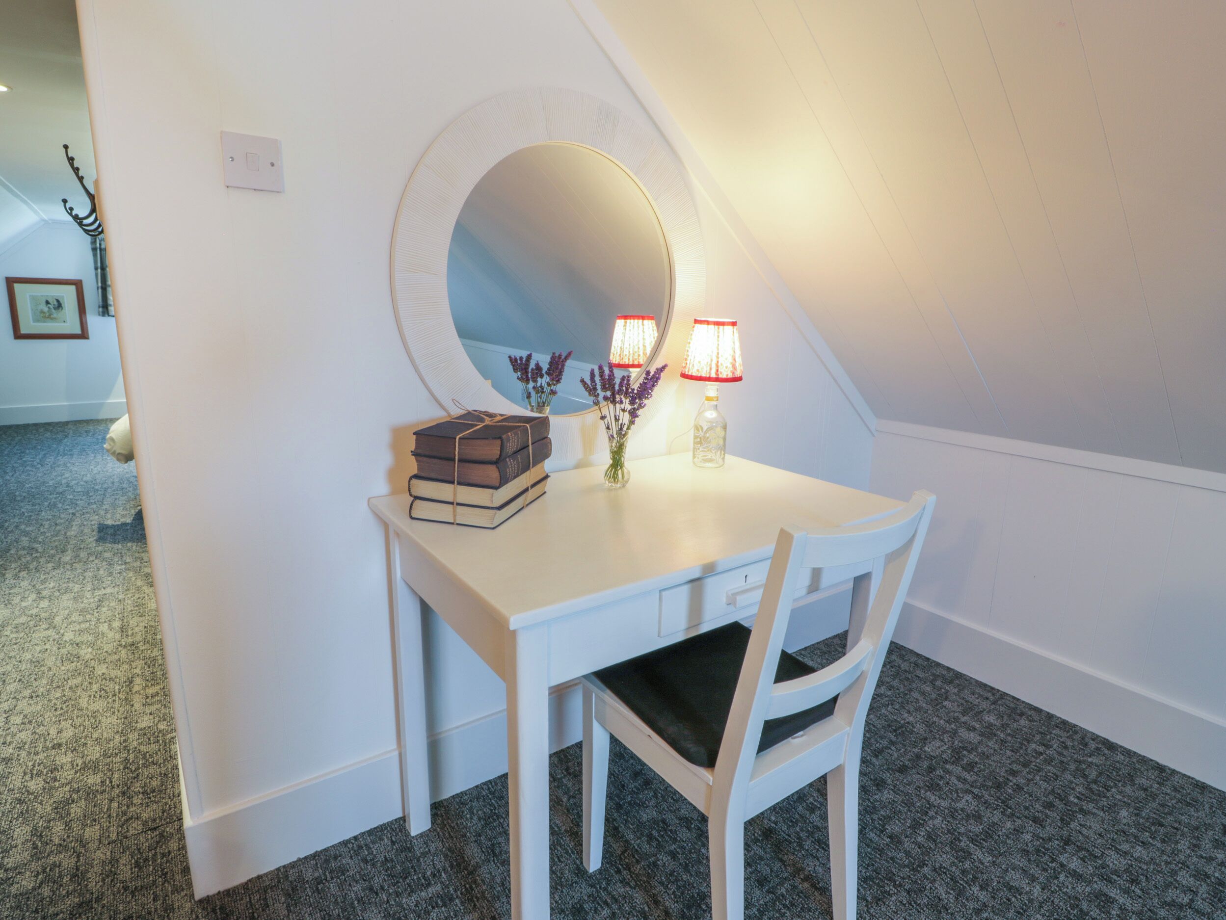A white desk with a mirror, books, and a vase with flowers in a corner room.
