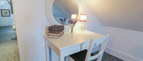 A white desk with a mirror, books, and a vase with flowers in a corner room.