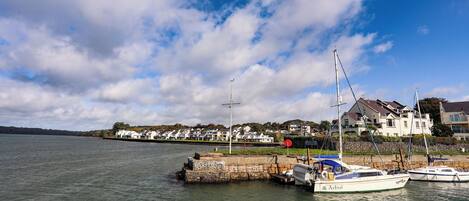A marina with boats docked, a stone jetty, and houses in the background.