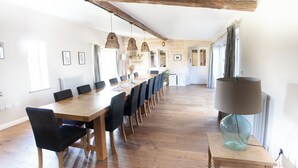 A spacious dining area with a long wooden table and chairs, a hanging light fixture, and a view of the kitchen in the background. - Hammonds Farmhouse (Stroud)