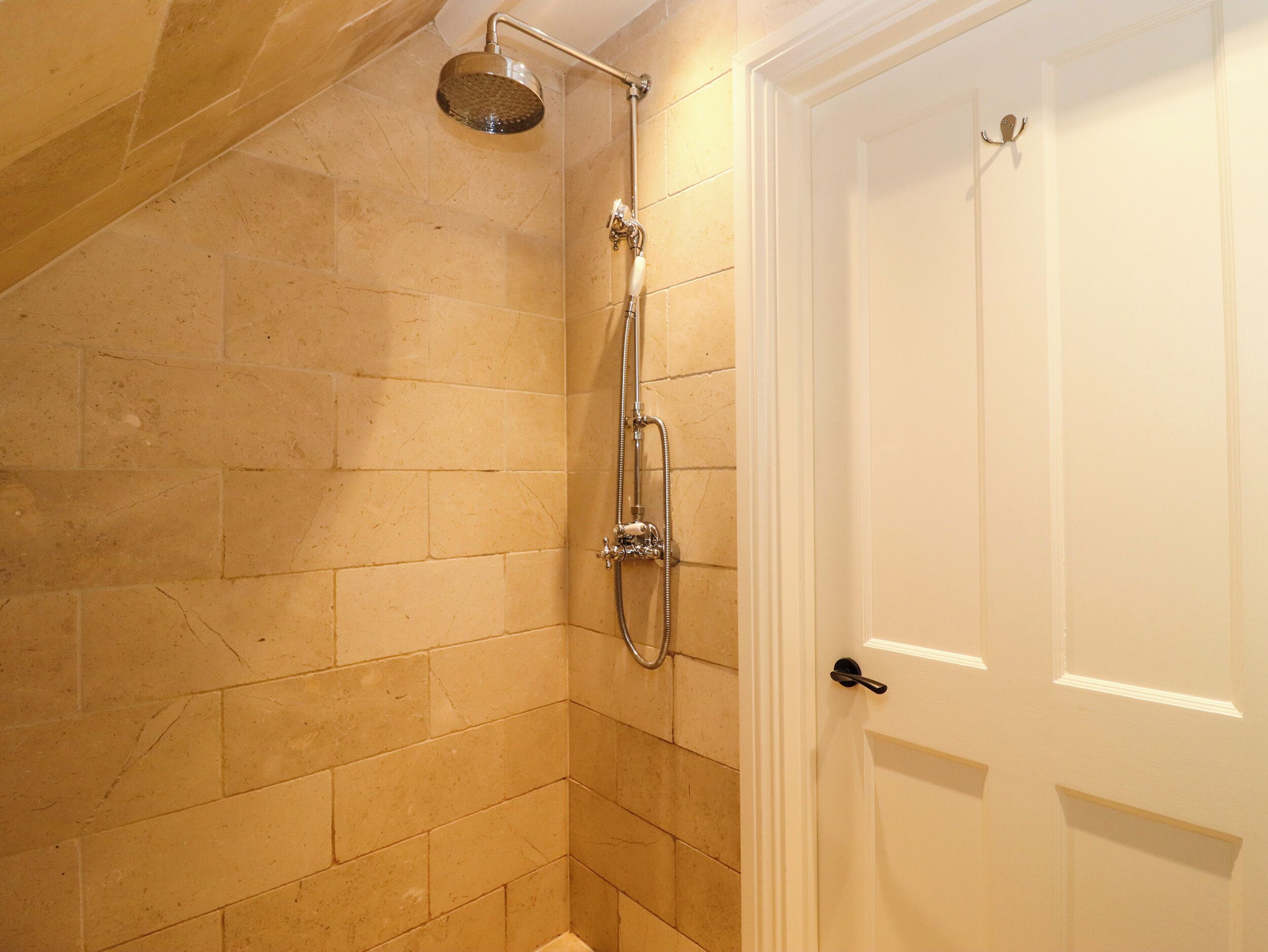 A shower with a handheld showerhead and a wall-mounted faucet, a white door with a black handle, and beige tiled walls.