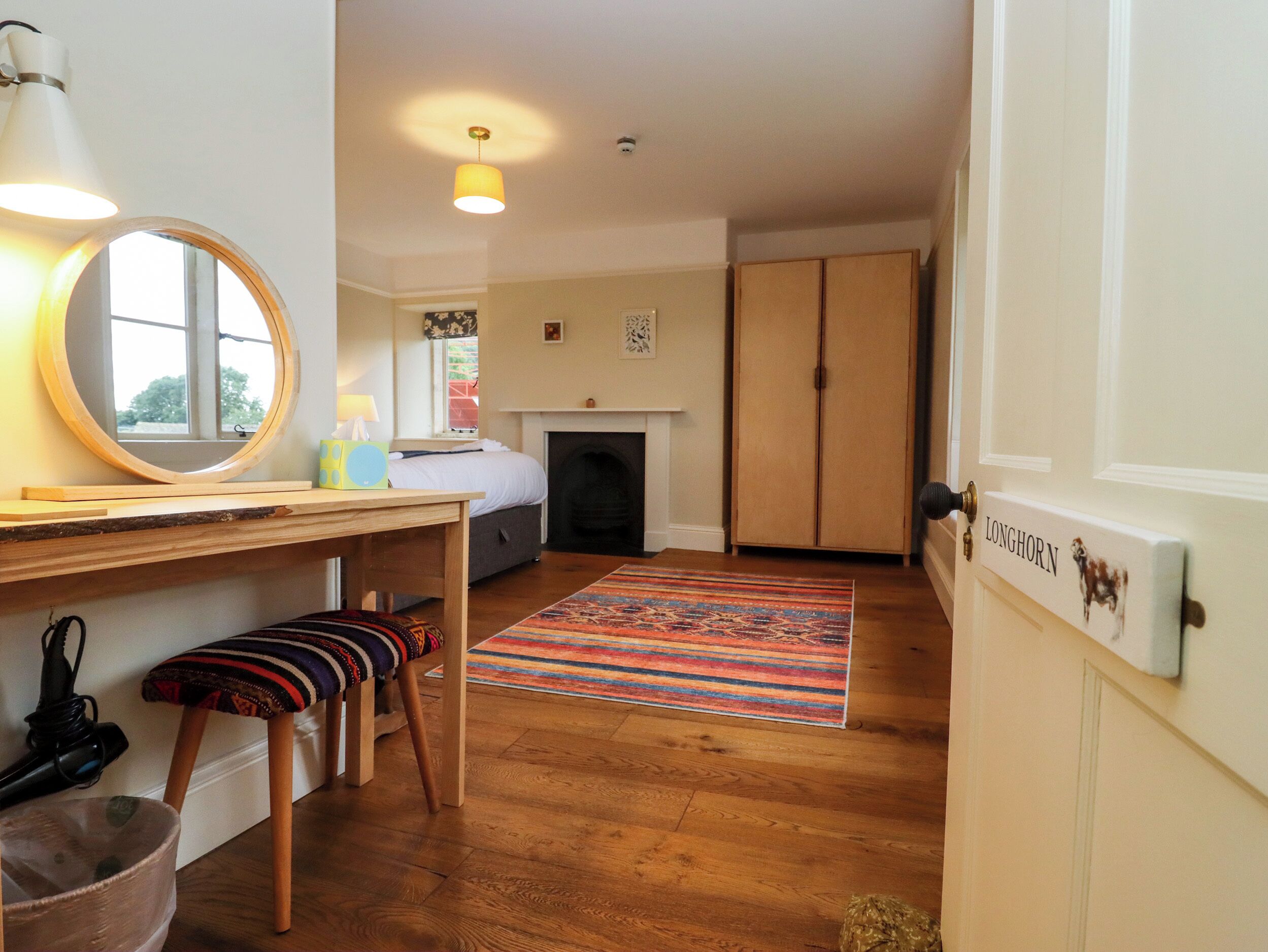 A cozy room with a wooden desk, a round mirror, a bed, a fireplace, and a colorful rug.