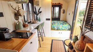 A compact kitchen with a wooden countertop, a kettle, a coffee machine, and a sink. There is a window with blinds and a bed with a colorful floral bedspread. - Herdwick Luxury Glamping (Retford)