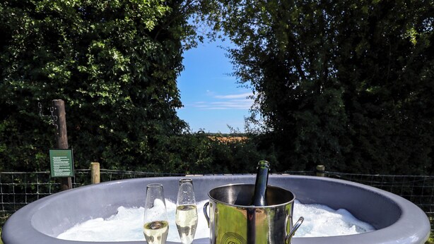 A hot tub with two glasses of champagne and a bottle, set on a wooden deck with a sign in the background. - Herdwick Luxury Glamping (Retford)