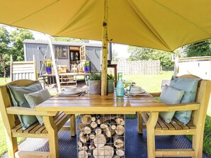 A wooden dining table with chairs, a yellow canopy, and a view of a corrugated metal building in the background. - Herdwick Luxury Glamping (Retford)