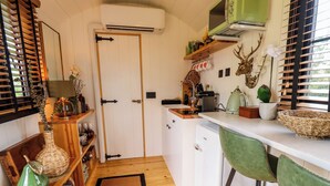 A compact kitchen with a white refrigerator, a green chair, and a wooden shelf with various kitchen items. - Herdwick Luxury Glamping (Retford)
