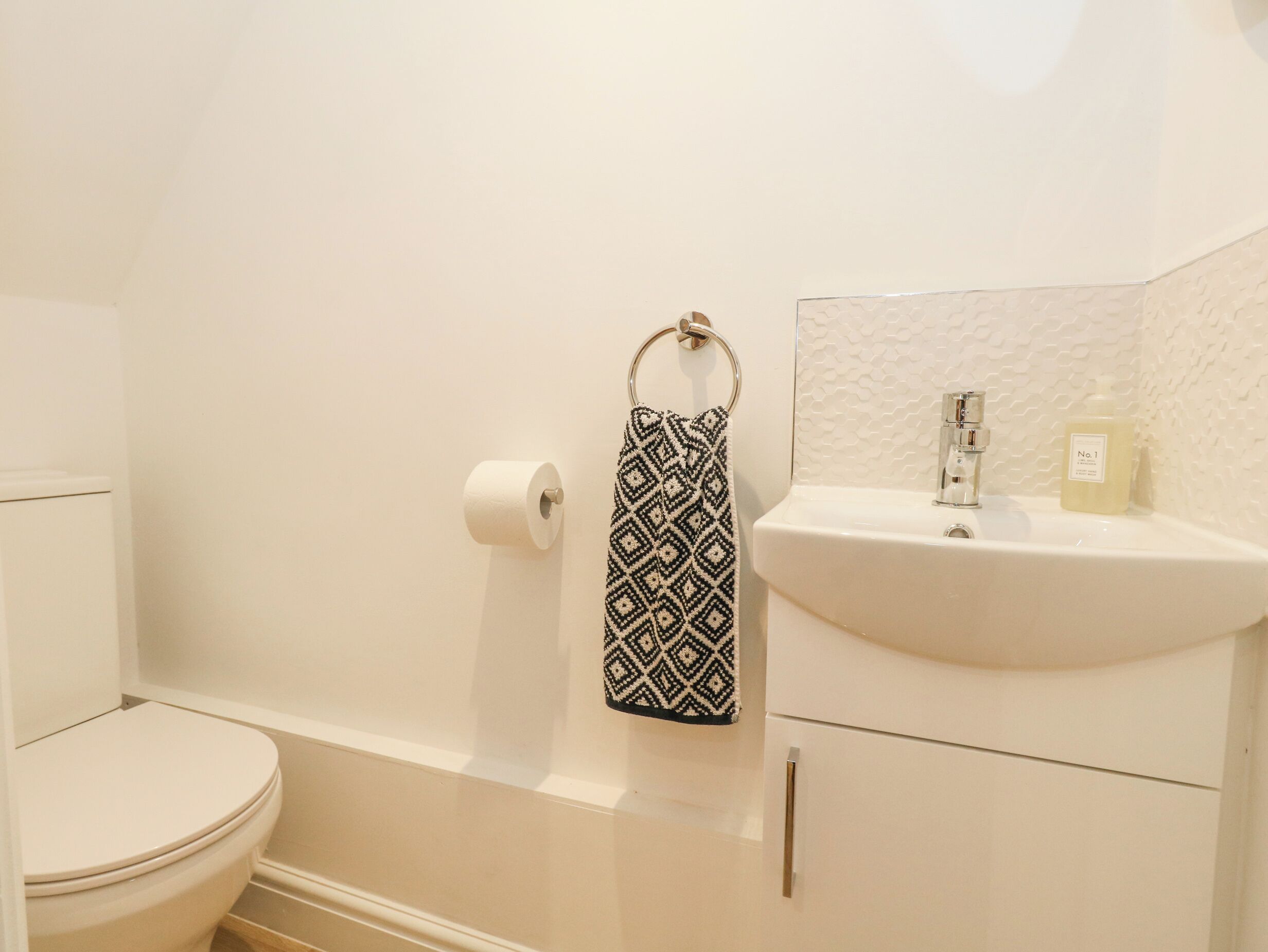 A bathroom with a toilet, a roll of toilet paper, a patterned towel hanging on a ring, and a sink with a soap dispenser.