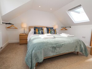 Une chambre avec un plafond en pente, un puits de lumière, un lit recouvert d’une couverture verte, deux oreillers, une table de chevet avec une lampe et une commode en bois.