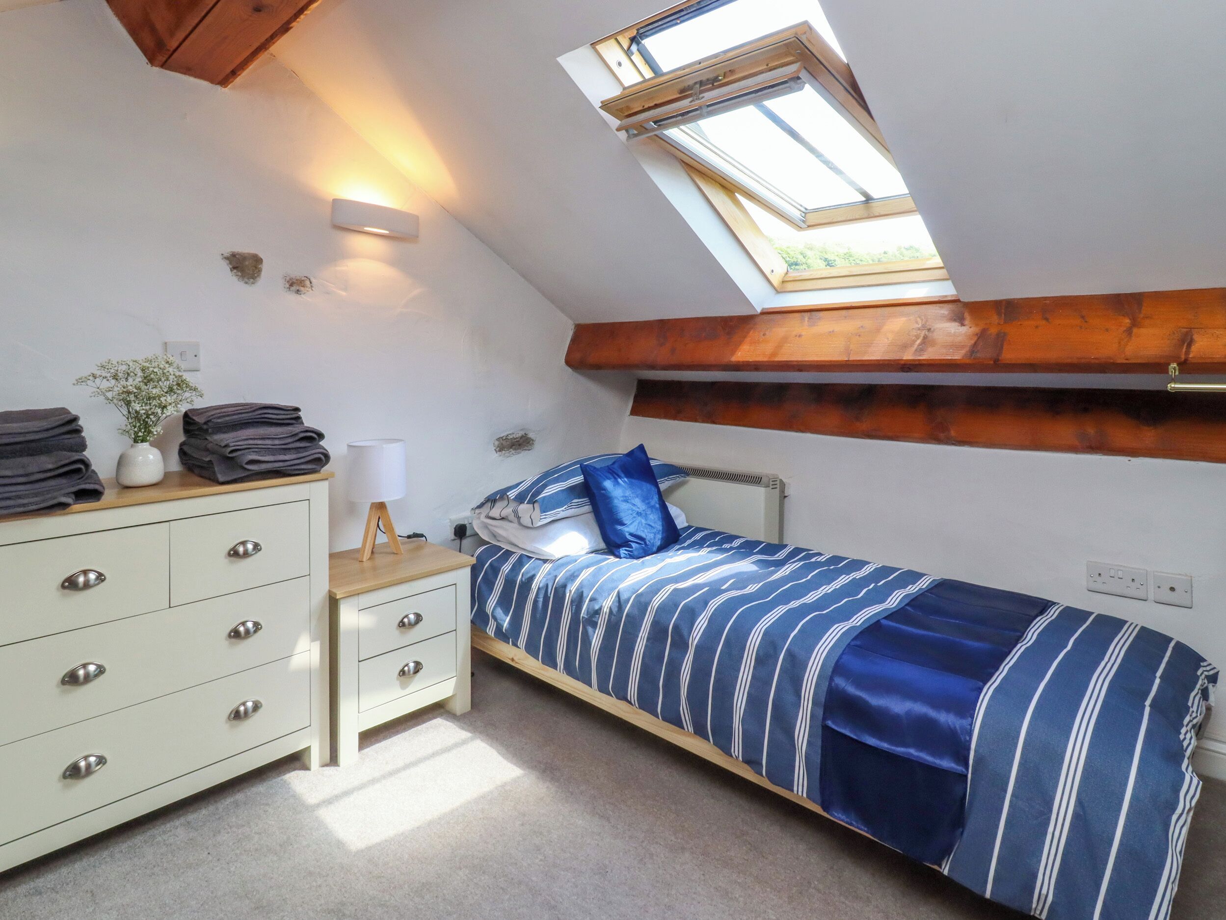 Une chambre avec un plafond en pente, une lucarne, un lit avec une literie à rayures, une table de chevet avec une lampe, une commode avec des tiroirs et une petite plante.
