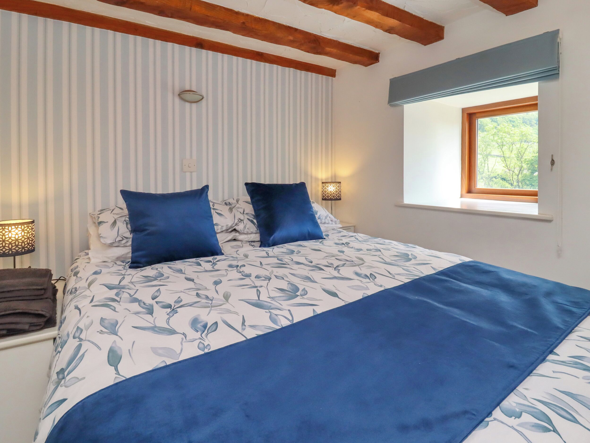 Un dormitorio con una cama, dos almohadas azules, una lámpara de noche, una ventana con vistas a árboles y una pared a rayas.