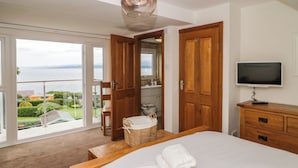 Une chambre avec un grand lit, une commode en bois, une télévision et une vue sur la mer, visible à travers une porte-fenêtre coulissante.