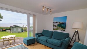 Ein Wohnzimmer mit einem türkisfarbenen Sofa, einem kleinen Tisch und einer Lampe. Ein großes Fenster bietet Blick ins Freie.