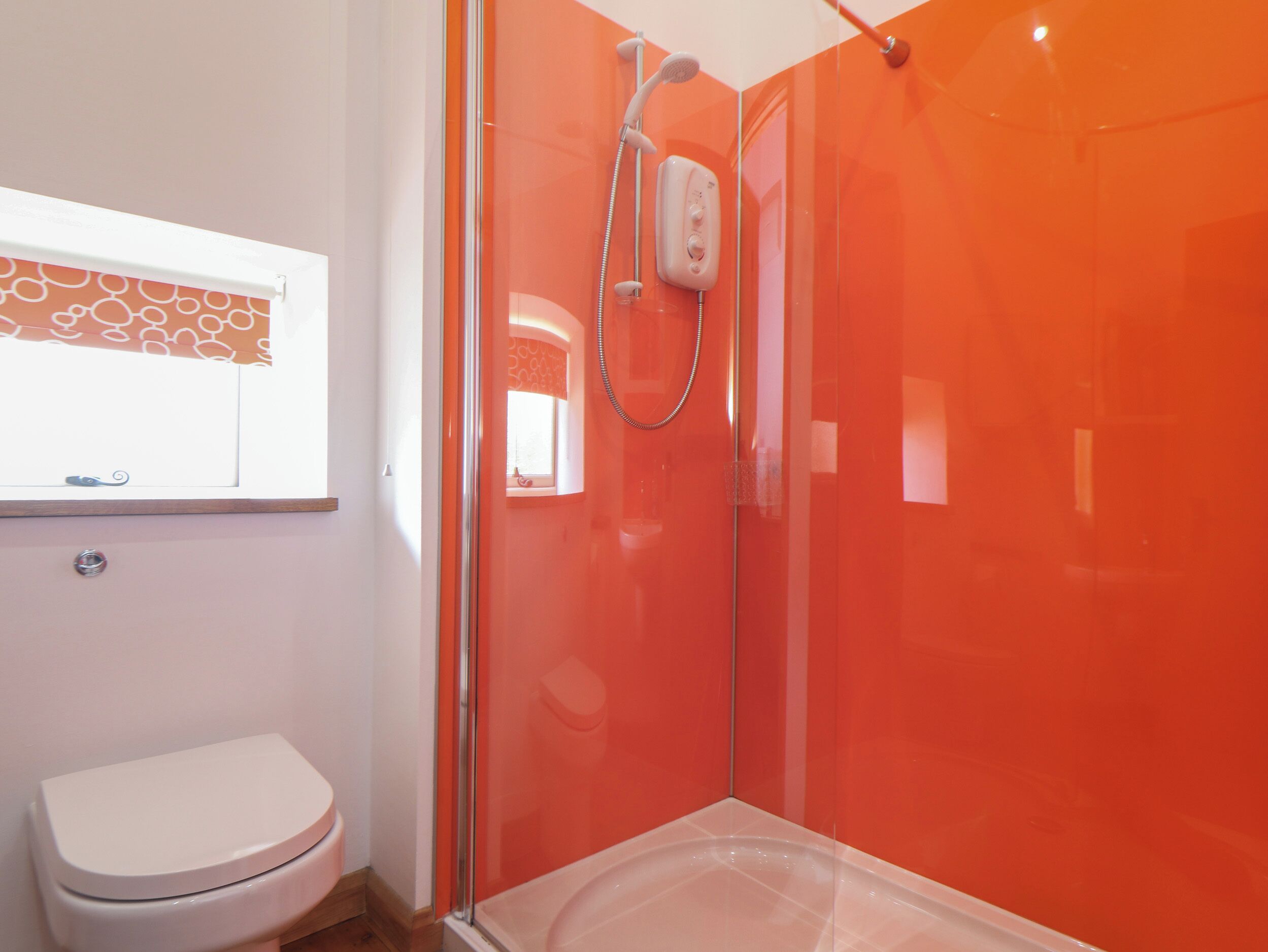 A modern bathroom with a glass shower enclosure, a white toilet, and a window with a patterned curtain.