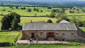 A stone building with a red door and windows, surrounded by green fields and hay bales, with a view of distant hills. - The Hayloft (Appleby-in-Westmorland)