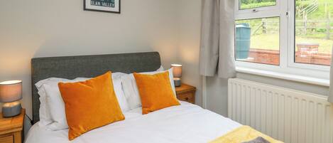 A neatly made bed with white and orange bedding, a framed picture on the wall, and a window with a view of greenery.