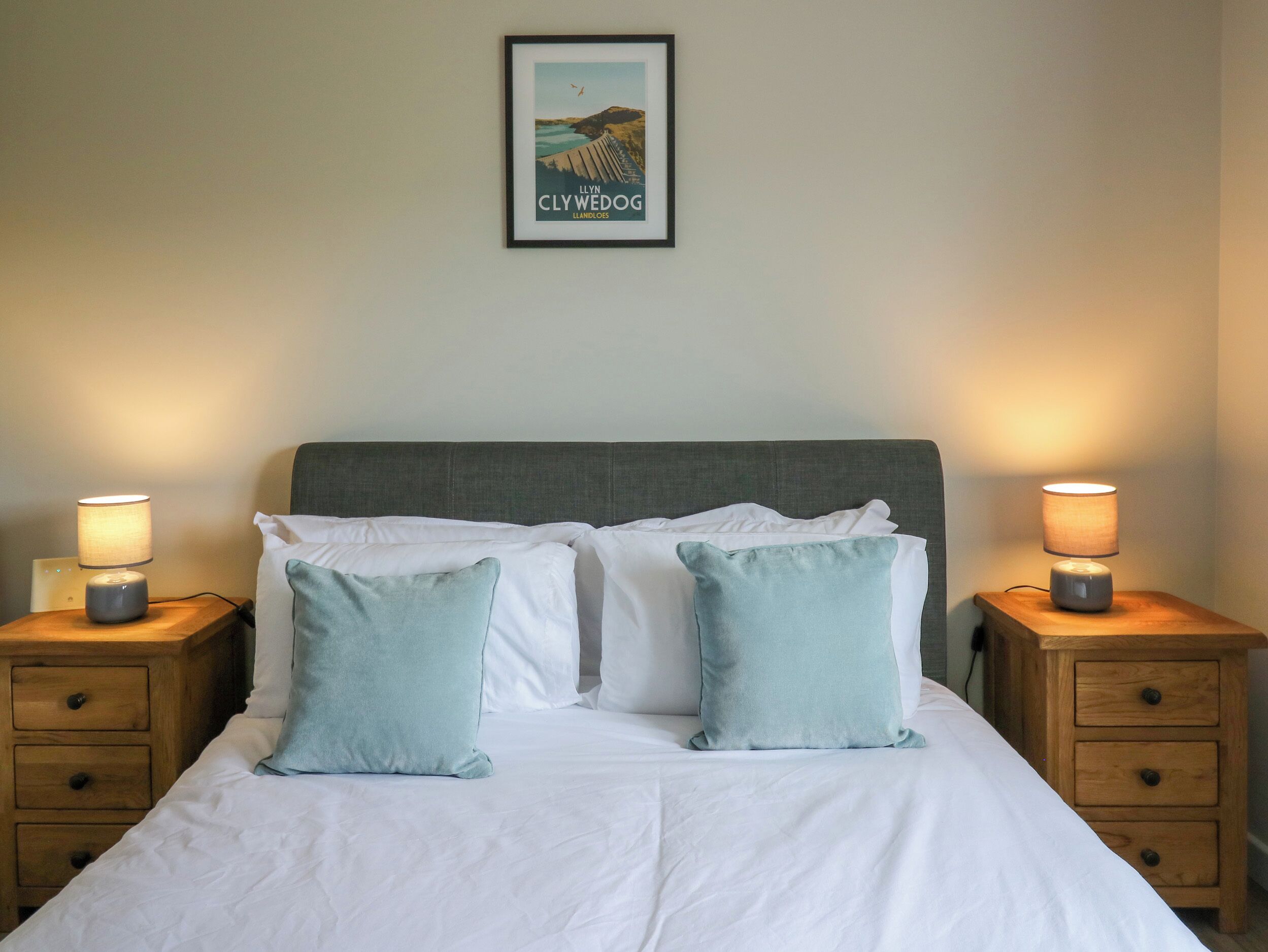 A neatly made bed with two pillows, a framed picture on the wall, and two bedside lamps on wooden nightstands.
