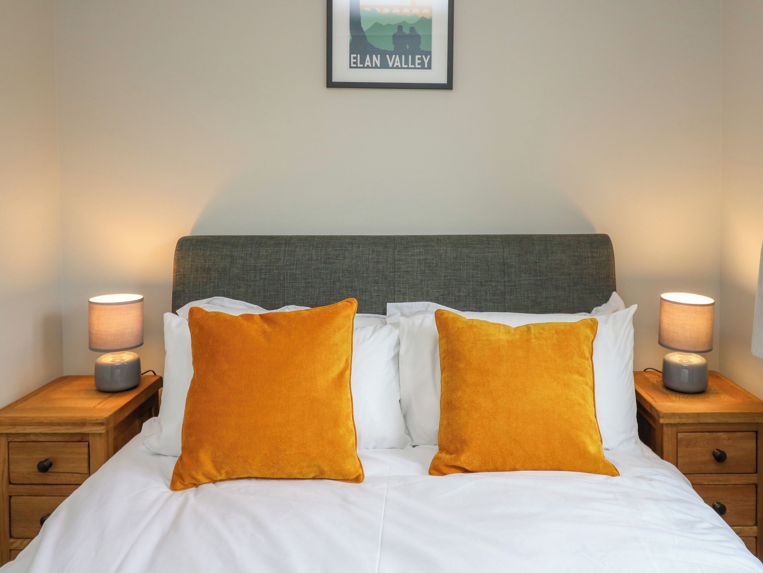 Een net opgemaakt bed met witte lakens en twee mosterdgele kussens, aan weerszijden geflankeerd door nachtkastjes met lampen, in een kamer met een lijstwerkfoto van Elan Valley aan de muur.