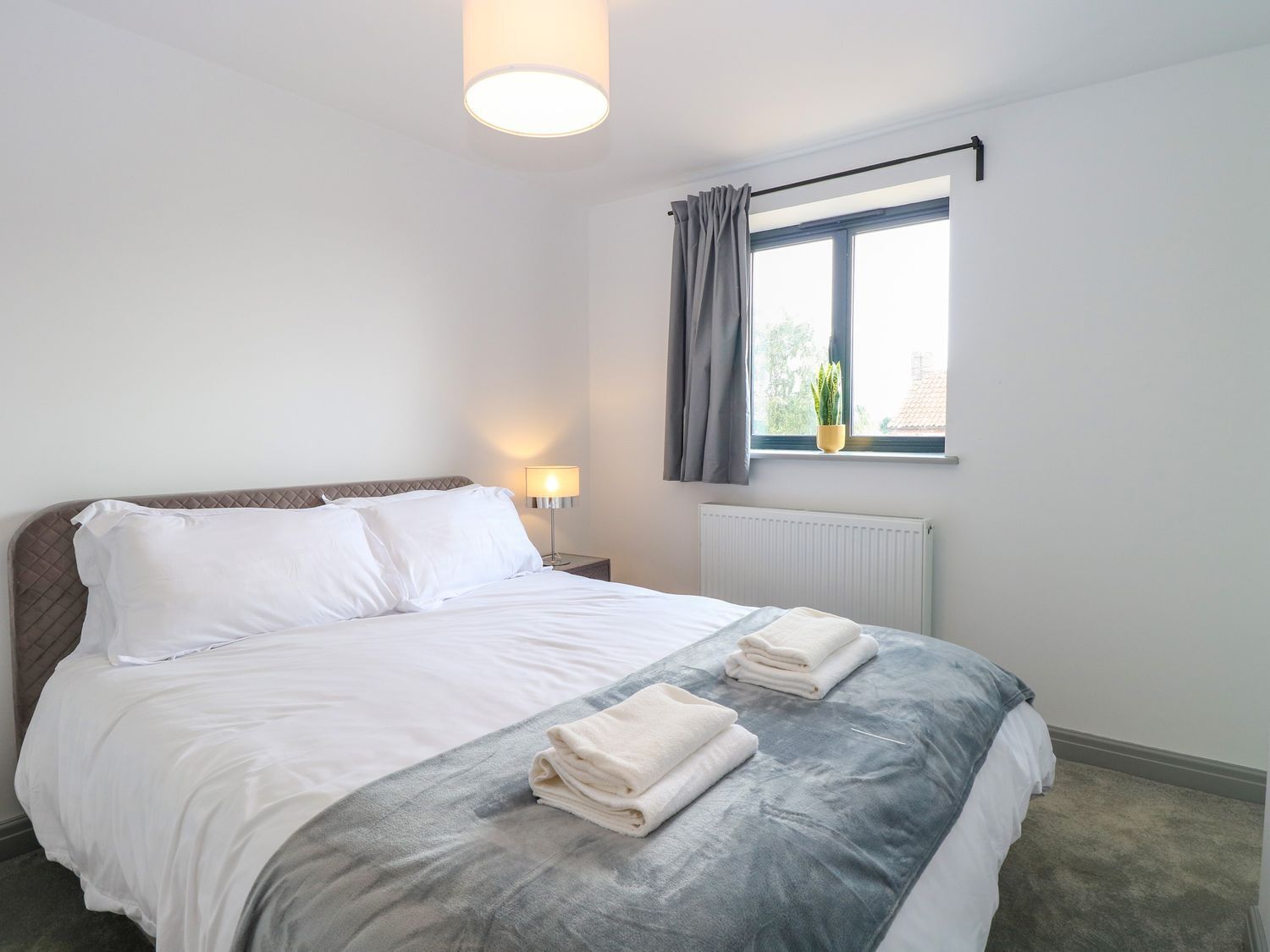 A neatly made bed with white linens and a grey blanket, a bedside lamp, a window with curtains, and a radiator.