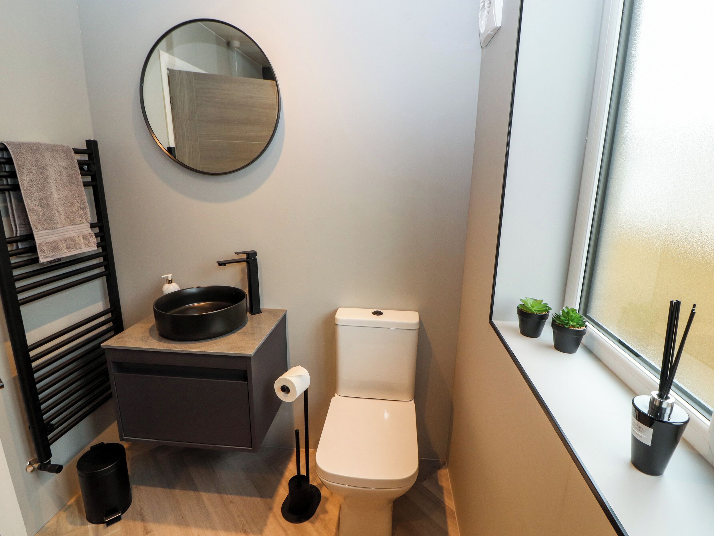 A compact bathroom with a toilet, sink, and mirror. There are small potted plants on the windowsill.