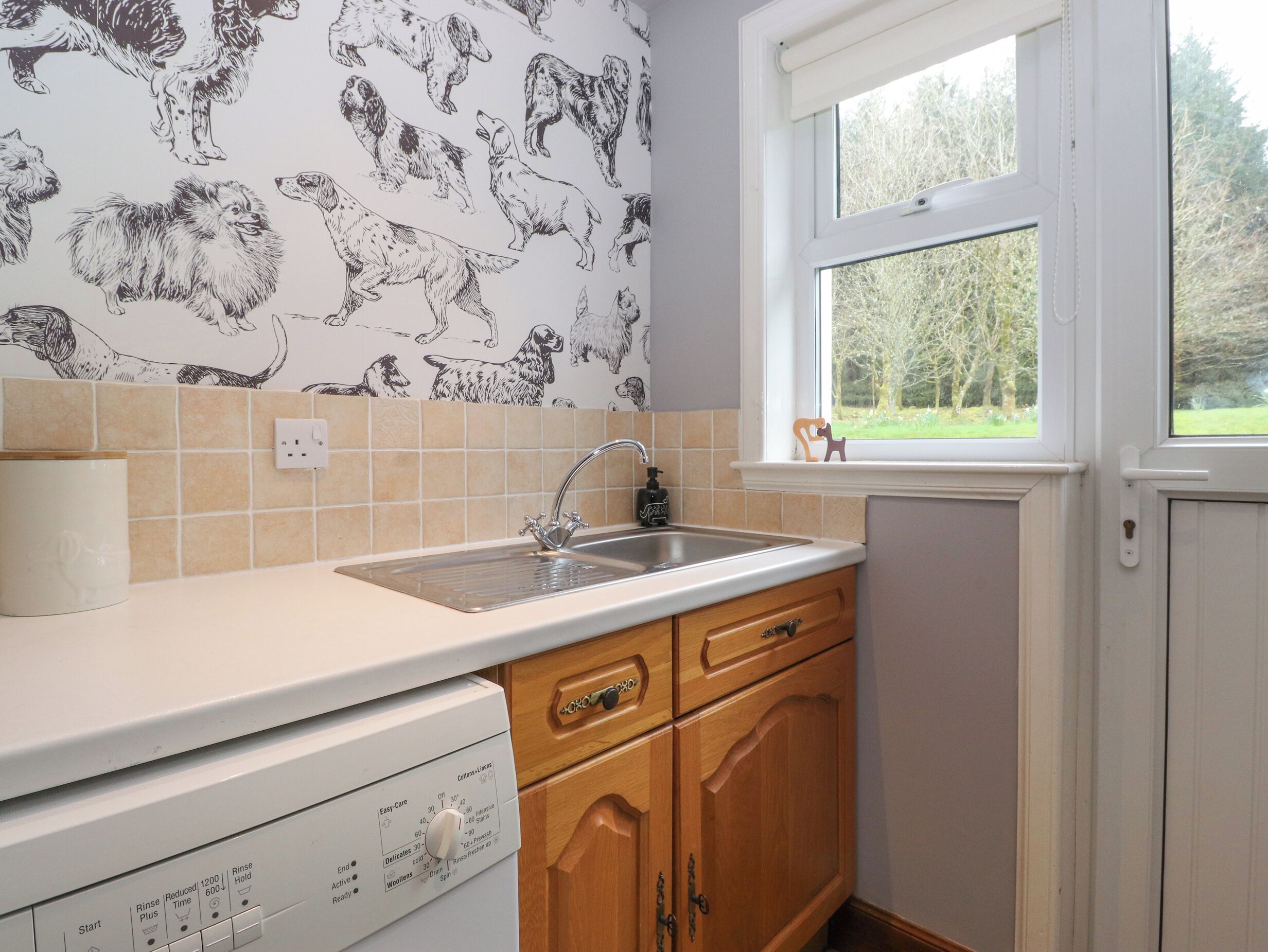 A kitchen with a sink, wooden cabinets, a dishwasher, and a window with a view of trees.
