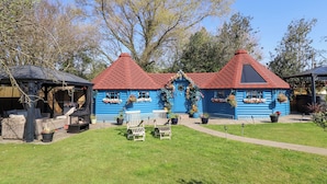 A blue wooden house with a red roof, surrounded by a grassy lawn and trees. - Applejacks Orchard (Holywell)