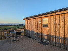 A wooden building with a door and window, a deck with chairs, and a view of a green landscape.