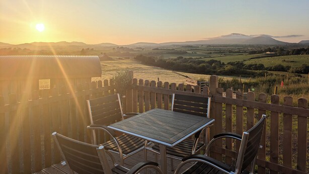 A wooden deck with a table and chairs overlooking a scenic valley with a mountain in the distance. - Hafan Gelli Sheperds Hut - Enlli (Pwllheli)