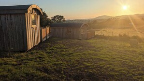 A wooden barn with a fenced area and a small shed, set against a backdrop of a sunset over a grassy field. - Hafan Gelli Sheperds Hut - Enlli (Pwllheli)