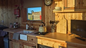 A wooden cabin interior with a kitchen area, including a sink, stove, and a window with a view of a wooden fence. - Hafan Gelli Sheperds Hut - Enlli (Pwllheli)