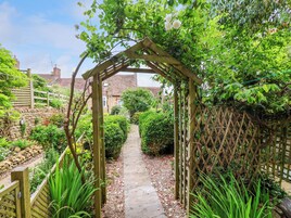 Uma trilha no jardim leva a um arco de madeira, cercada por vegetação exuberante e uma parede de pedra.