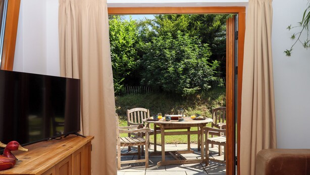 Habitación con televisor sobre un mueble de madera, una mesa con sillas y un ventanal corredizo que da a un espacio exterior con una mesa para dos.