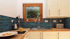 Una cocina con azulejos azul verdosos, una ventana con vistas a follaje, un lavamanos, un hervidor y un plato con cruasanes.