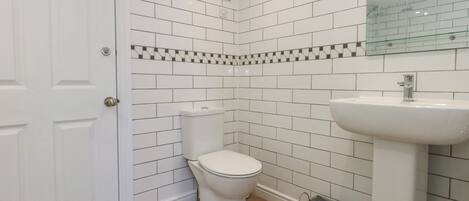 A bathroom with white subway tiles, a white toilet, a sink, and a small laundry basket.