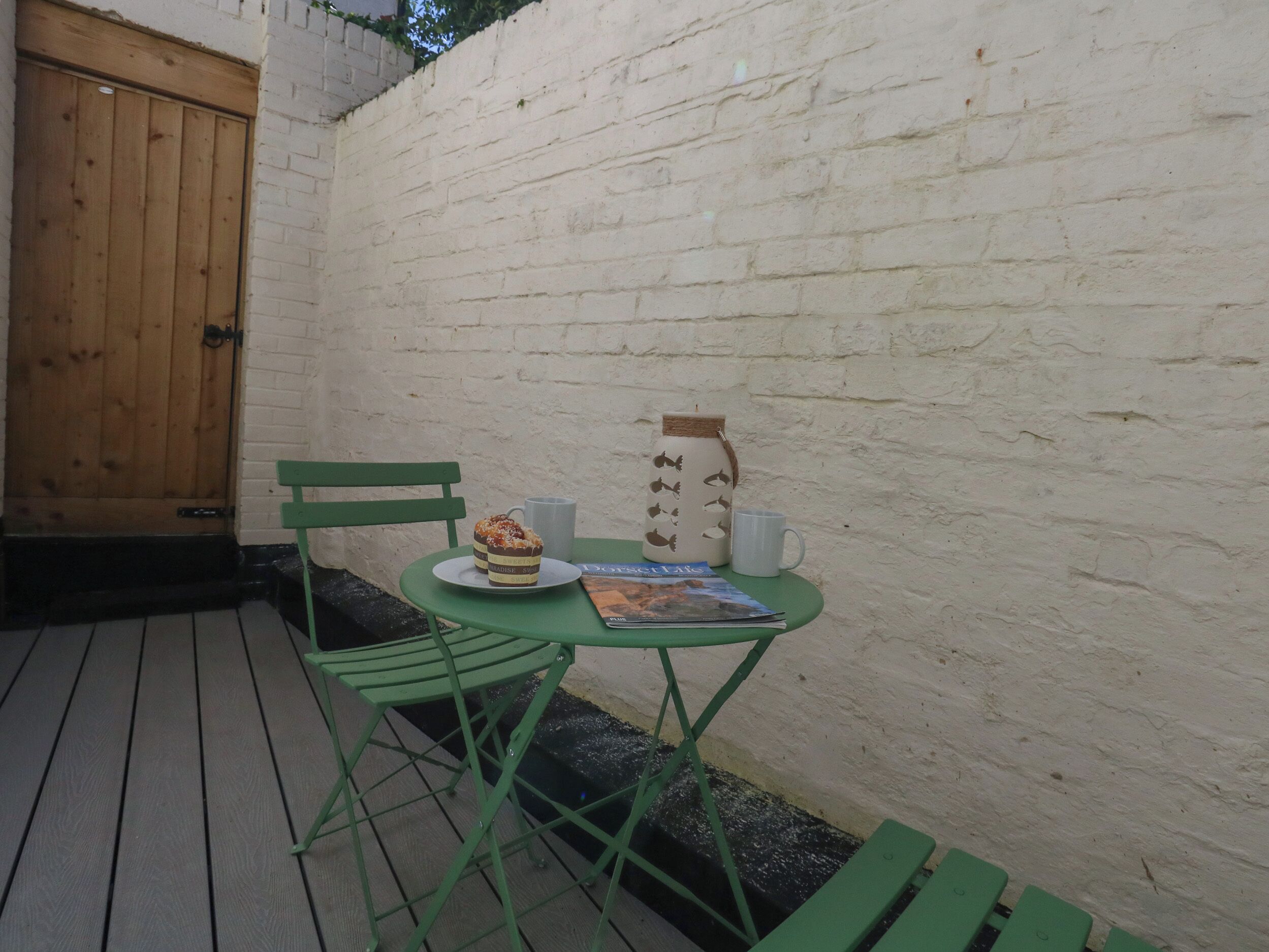 Un pequeño espacio al aire libre con una mesa y sillas de metal verde, una puerta de madera y un muro de ladrillo blanco.