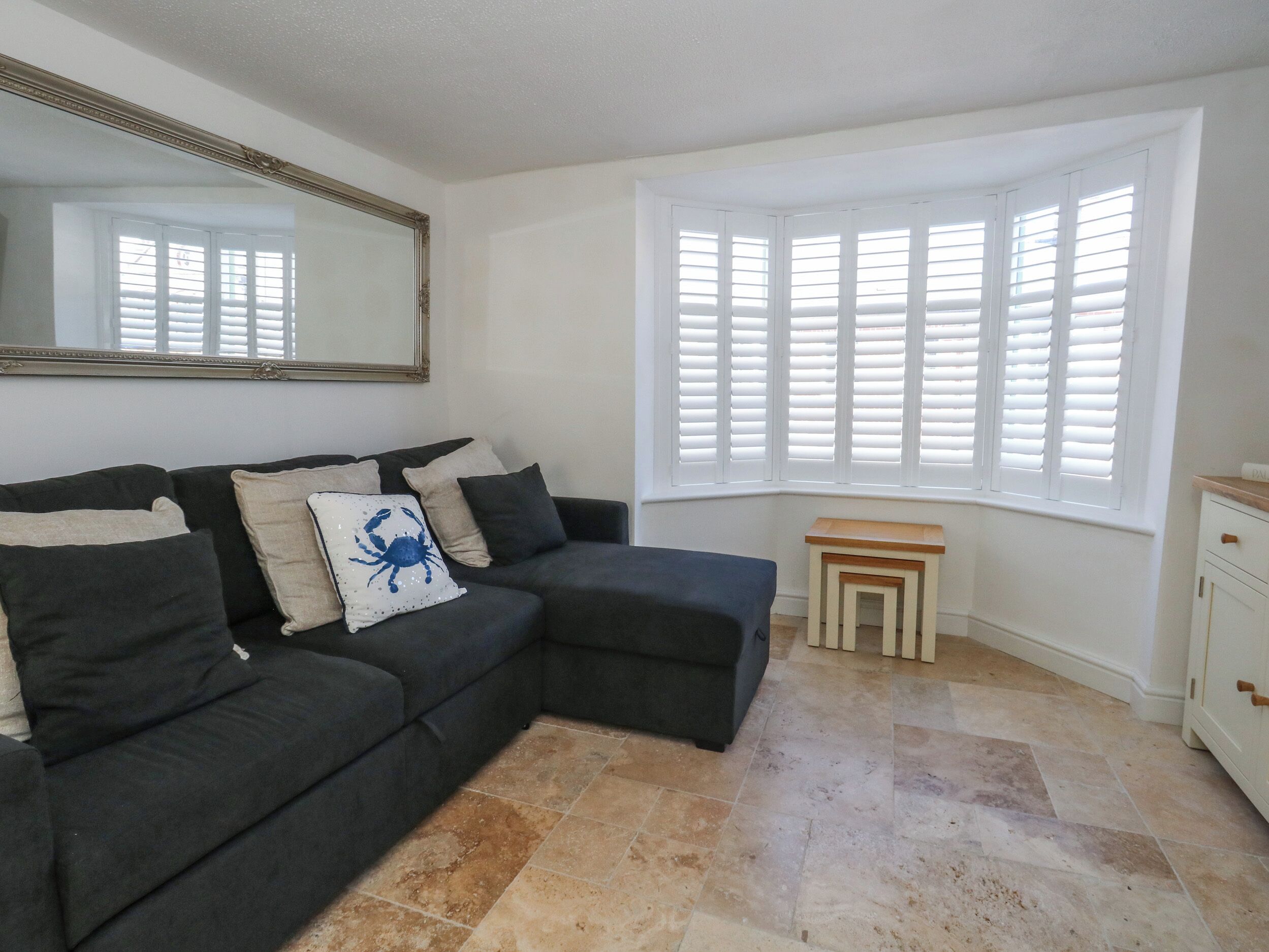 A room with a dark-colored sofa, a small wooden side table, a window with white shutters, and a mirror on the wall.