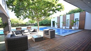 Una terraza de madera con muebles de mimbre, una piscina y un edificio moderno con grandes ventanales.