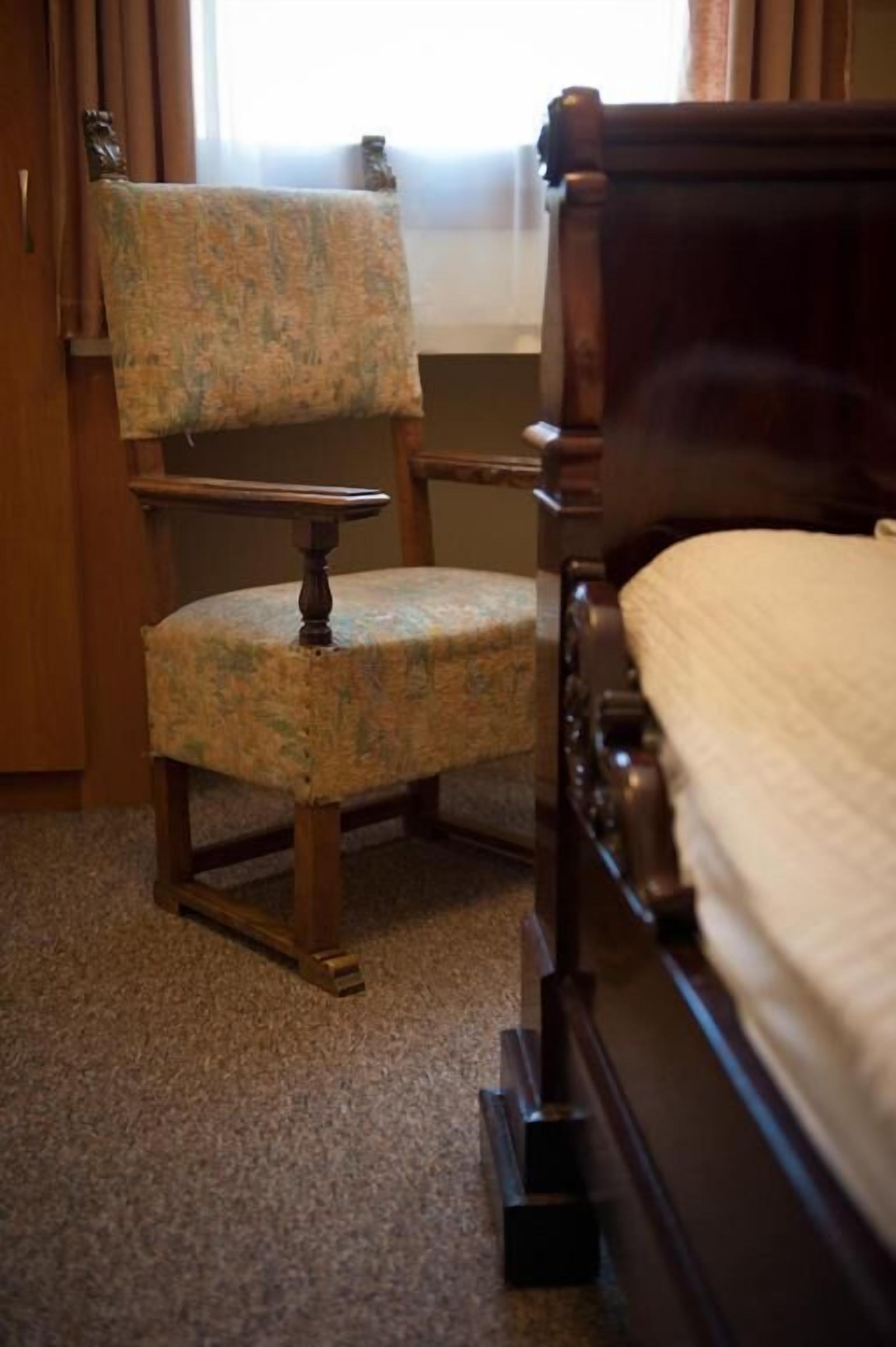A bedroom with a wooden bed, a floral-patterned chair, and a carpeted floor.