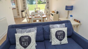 A cozy living room with a blue sofa, two pillows with 'HAPPY PLACE' text, a dining area with a table set, and a view of a garden through a glass door. - Whitfield P5 (Yarmouth)