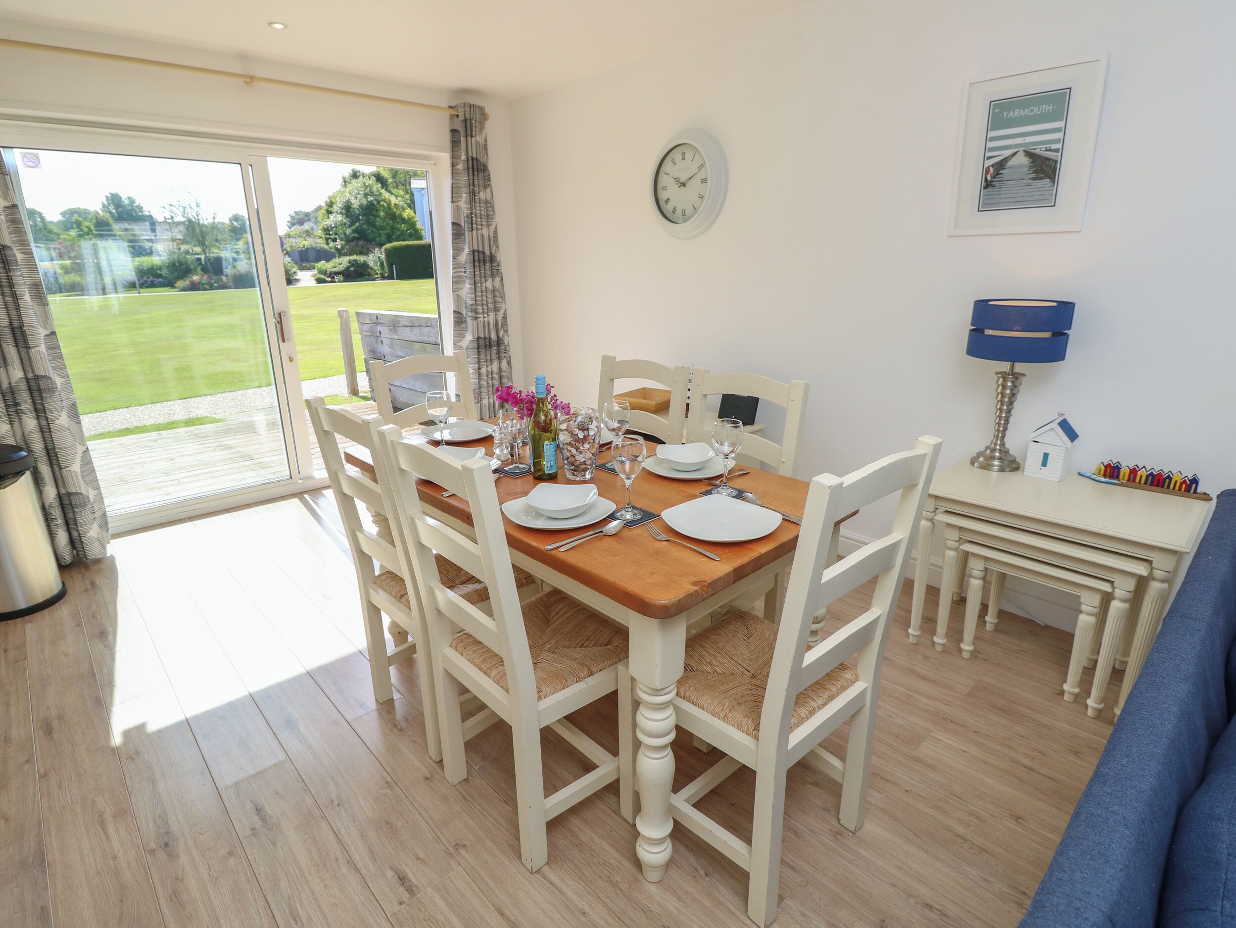 A dining area with a wooden table set for four, white chairs, a clock on the wall, and a view of a garden through a glass door.