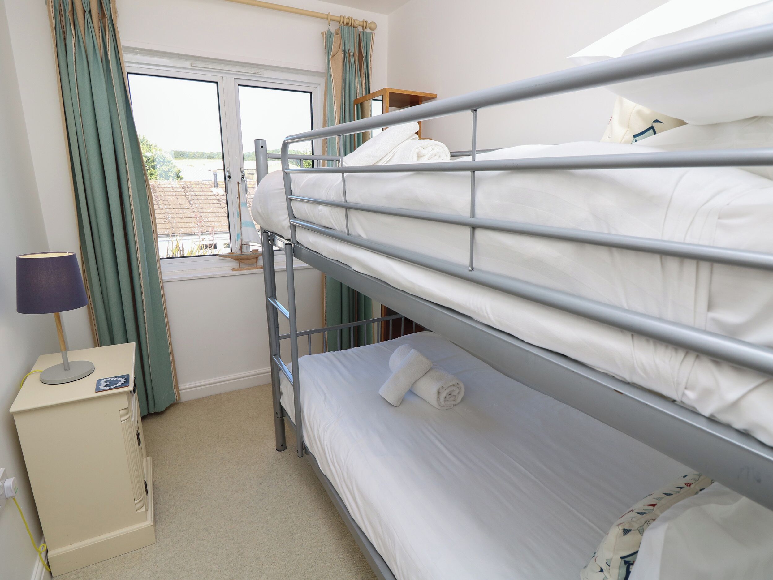 A bunk bed room with a window, curtains, a bedside table, and a lamp.