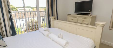 A neatly made bed with rolled towels, a television on a dresser, and a balcony with a view of the countryside.