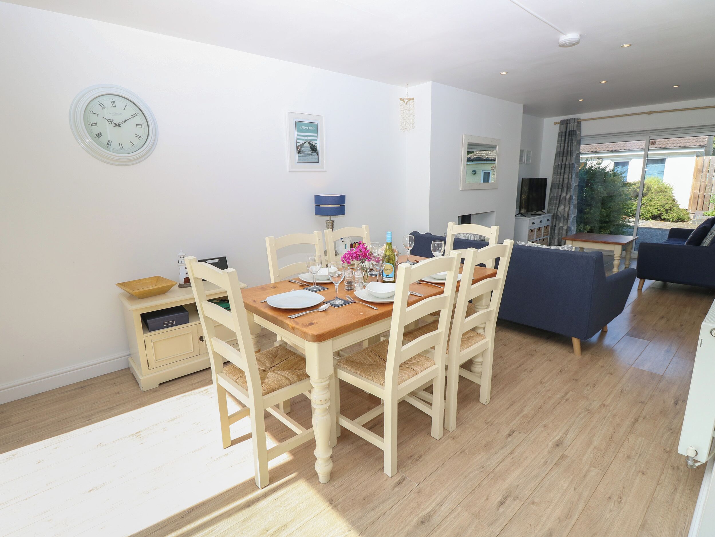 A dining area with a wooden table set for four, white chairs, and a clock on the wall.