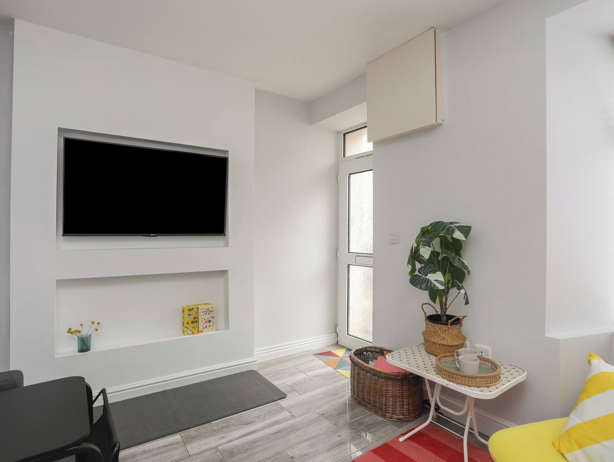 A modern living room with a flat-screen TV, a dining table, a yellow chair, a small plant, and a wicker basket.
