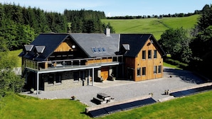 Een modern huis met een ruim terras, zonnepanelen en een prachtig uitzicht over een groen veld en bos.