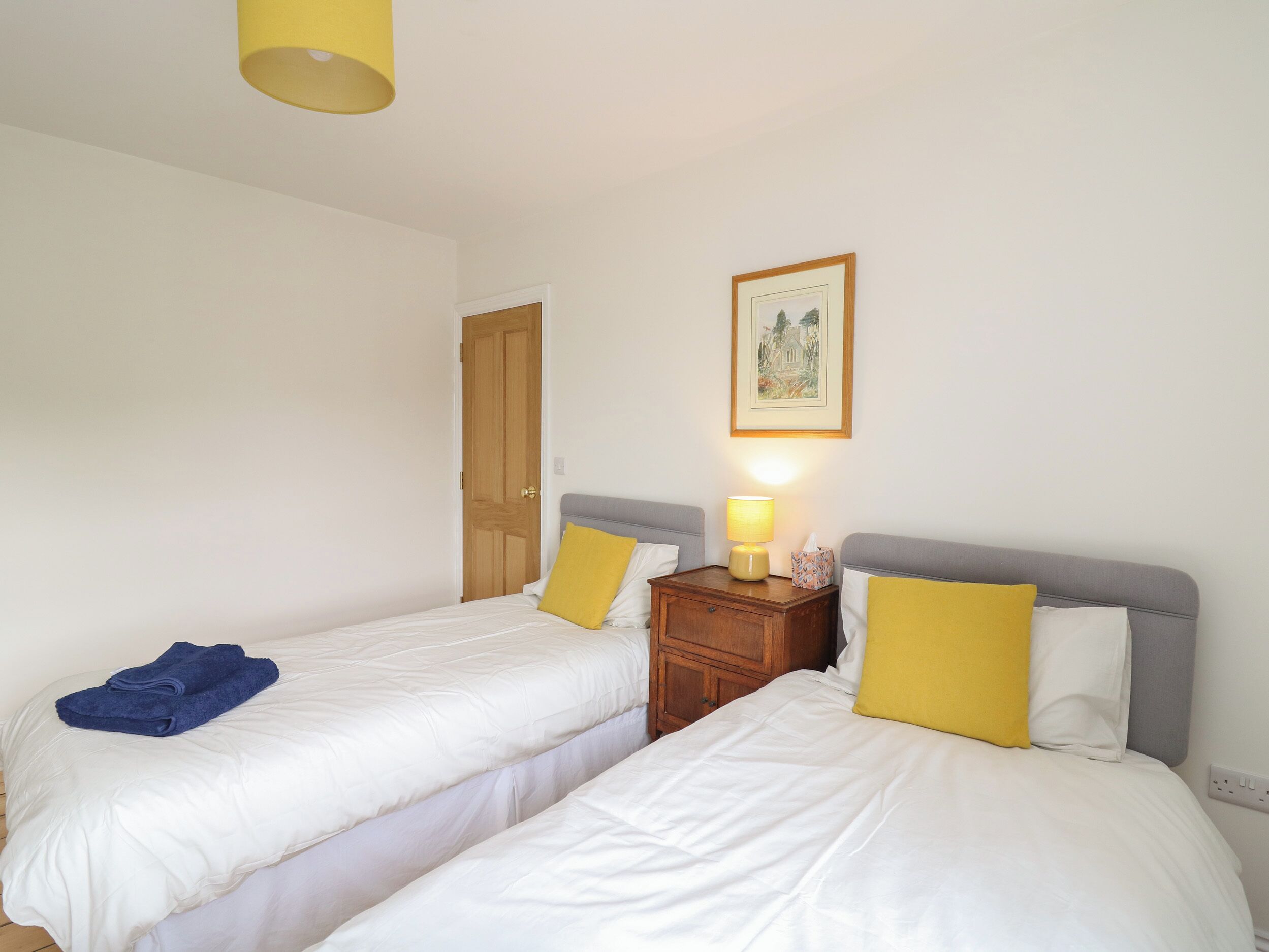 A hotel room with two beds, a wooden nightstand, a lamp, and a framed picture on the wall.
