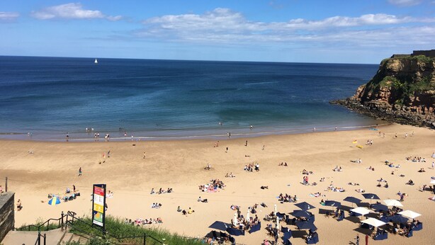 A beach with people sunbathing and swimming, a sailboat in the distance, and a rocky cliff with vegetation. - Limpet Lodge (North Shields)