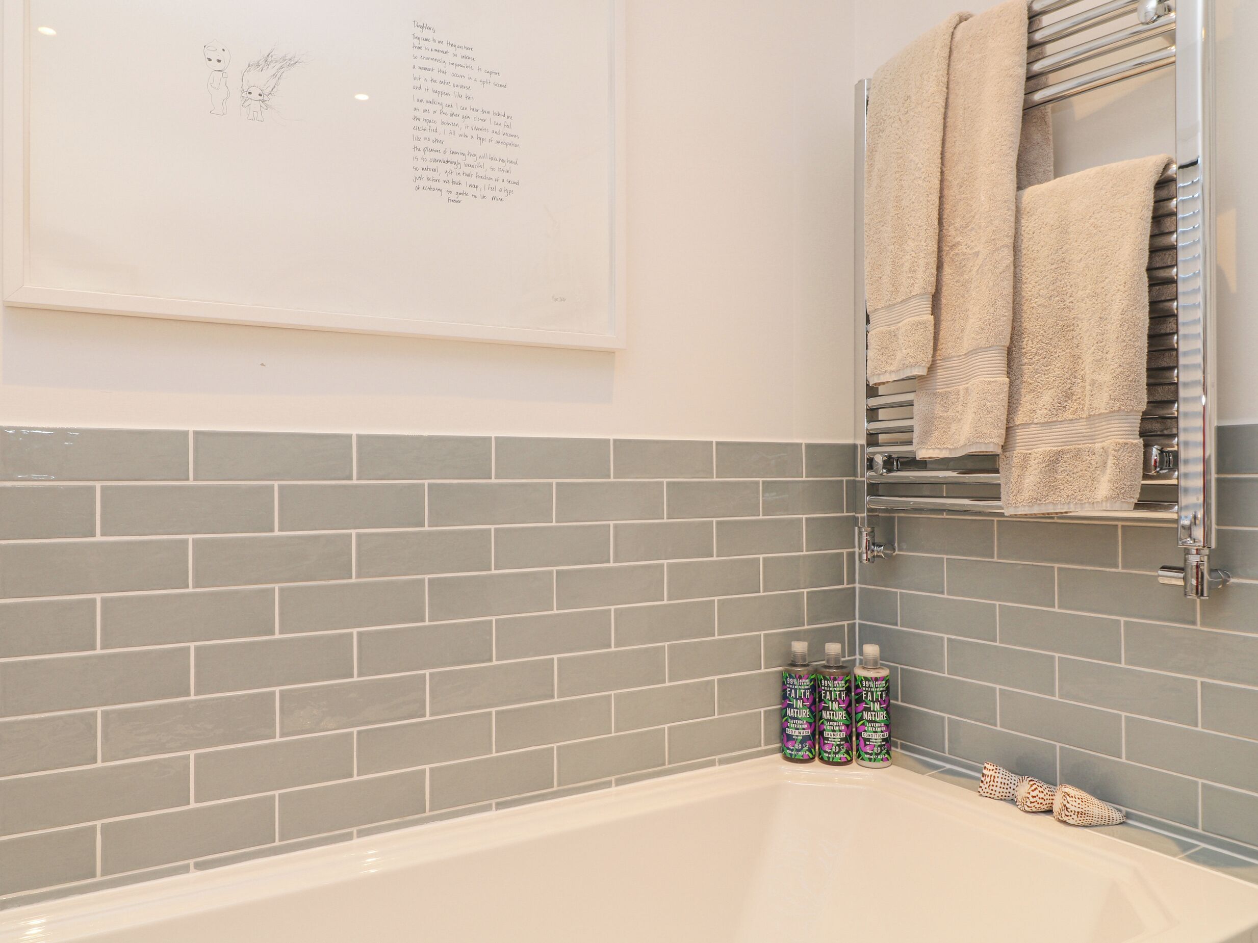 A bathroom with grey tiled walls, a white bathtub, and a towel rack holding two towels.